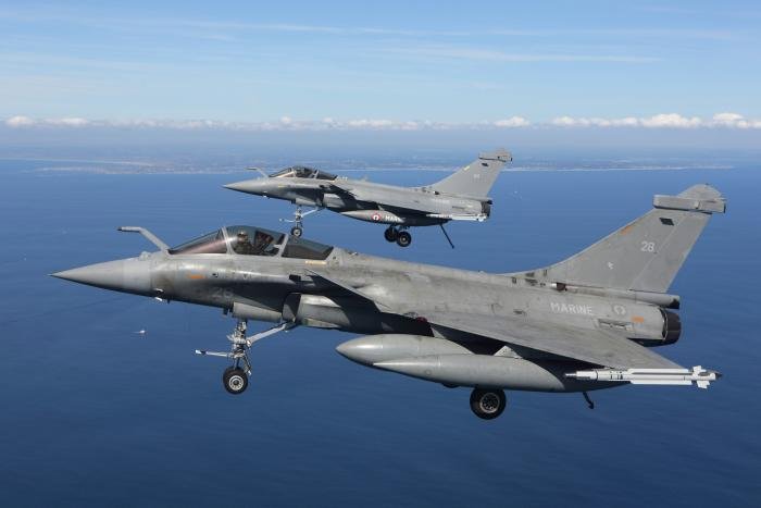 Two Flottille 11F Rafale M fighters fly with the landing gear extended to show their massive front landing legs and their launch bars. Their tail hooks have also been lowered, in French Navy tradition.